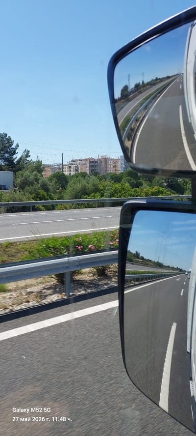 The image depicts a view from a moving vehicle on a highway in l'Ametlla de Mar, Spain, as inferred from the reflection in the side mirrors. The road surface is asphalt with white lane markings. A metal guardrail runs along the side of the road, adjacent to green vegetation and some pink flowers. In the left side mirror, the reflection shows the same highway continuing onward, curving slightly with additional buildings and greenery visible further in the distance. The sky is clear and blue. The image includes timestamp data indicating it was taken with a Galaxy M52 5G on May 27, 2026, at 11:48.