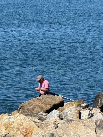 A man fishes from a rocky shore, with the deep blue ocean stretching out behind him. He is wearing a pink polo shirt and a camouflage baseball cap, and is focused on something in his hands. Two fishing rods are visible, one extending into the water and another lying on the rocks. The sun is bright, casting sharp shadows on the rocks, suggesting it is midday. The location is the Gafanha da Nazaré, Portugal, a coastal area known for its fishing traditions and rugged coastline.
