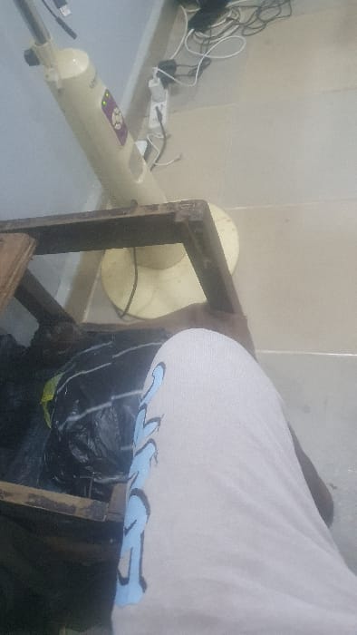 An indoor scene is captured from a low perspective, showing a human leg in the foreground, wearing light-colored pants with blue and black graphic lettering. Behind the leg and positioned on a light-colored tiled floor, a dark brown wooden table or stool frame is visible, beneath which rests a crumpled black plastic bag. In the background, an operational beige tower fan, identified by illuminated green indicator lights on its purple control panel, stands on the floor. Adjacent to the fan, against a light-colored wall, is an electrical setup including a power strip or wall socket with multiple black and white cables and adapters connected. The specific location within Danbare, Nigeria, is not discernible from the visual evidence.