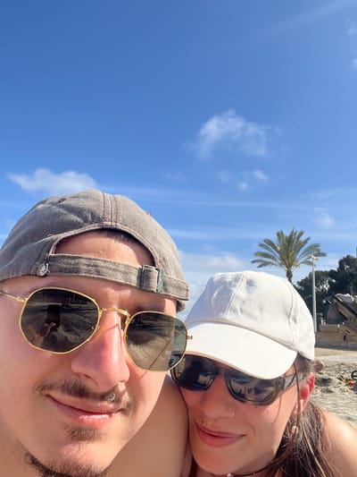 A selfie was taken on a sunny day at a beach in Peguera, Spain. The photo features two individuals. In the foreground is a person wearing sunglasses and a grey baseball cap. Behind this person and slightly above is another person wearing sunglasses and a white baseball cap. A palm tree is visible in the background. The sky is clear with some scattered clouds. Beach sand and other beach paraphernalia can also be seen in the background.