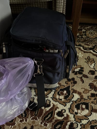 A dark blue backpack is positioned on a patterned carpet. A purple plastic bag is partially covering the front of the backpack, obscuring some of its details. The backpack has multiple zippers and straps visible. In the background, a dark wooden furniture leg and a portion of a patterned textile are discernible. The lighting suggests an indoor environment.