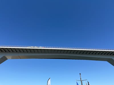 The image shows a wide, modern concrete bridge spanning across a clear blue sky. The underside of the bridge is visible, with its distinctive supporting structure. Along the top of the bridge, several cars are driving, suggesting it is a functional roadway. The cars appear to be in a line, with some white, blue, and red vehicles visible.

Below the bridge, a bright, cloudless blue sky dominates the lower portion of the frame. In the far lower right corner, a small portion of a mast with flags is visible, hinting at a marine setting. One flag, partially visible on the left, has some text on it, but it is too blurry to read. The lighting suggests it is daytime, likely a bright, sunny day given the clear sky and strong shadows cast by the bridge's structure. The overall impression is one of a clear, open, and modern environment, possibly a coastal or harbor area given the maritime hints.
