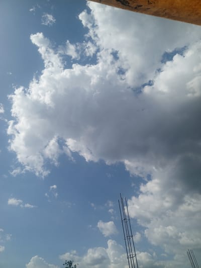 The image shows a view of the sky in Parroquia Ocumare, Venezuela. The sky is blue with numerous white clouds. A partially visible brown object is in the upper right corner of the image. In the foreground, there are vertical metal rebar structures extending upwards, likely part of a construction project. A small tree is visible at the bottom.
