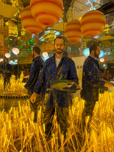 The image depicts a decorative display, likely for an exhibition or event, set in what appears to be an indoor location in Gongshu District, China. The primary subjects are multiple mannequin figures of Vincent van Gogh, each holding a painter's palette. These figures are positioned amongst an abundance of illuminated wheat stalks, casting a warm, golden glow over the scene.
Several large, spherical objects with orange and yellow stripes, resembling planets or large fruits, are also part of the display, positioned to the left of the figures. The background is a mirrored surface, reflecting the figures, wheat stalks, and spherical objects, creating a disorienting and immersive effect. The reflections also reveal glimpses of other decorative elements, including what appear to be colored lights and text in Chinese characters.
The overall impression is one of artistic representation and visual spectacle. The use of replicated figures and a thematic arrangement of elements suggests a curated experience, possibly related to art, nature, or celestial themes. The lighting, dominated by the warm tones of the wheat stalks and reflections, creates a festive or enchanting atmosphere, irrespective of the time of day or weather outside. No discernible emotions are conveyed by the static mannequin figures. Visible text includes "再见 Rè" and other characters, likely part of signage or exhibition labels.