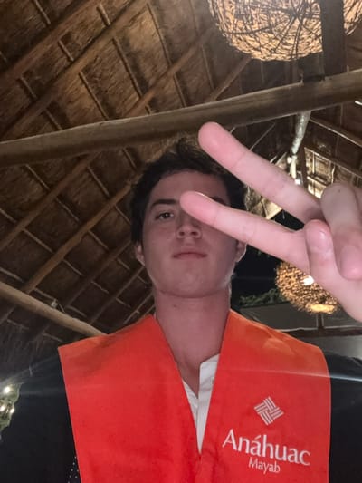 A vertically oriented, close-up photograph captures a young person making a peace sign with their hand, looking up towards the camera. The subject, likely male, has dark hair and a neutral to slightly smiling expression. They are wearing an orange vest or garment over a white shirt, with black attire visible underneath. Clearly visible on the orange garment is a white logo consisting of intertwined squares, and the text "Anáhuac Mayab."
The setting is an indoor or covered outdoor area, distinguished by a rustic, natural design. Above and behind the person is a ceiling made of tightly woven thatch or straw, supported by robust wooden beams and poles. To the left, a large, spherical light fixture crafted from woven material hangs, casting a warm glow. Additional, smaller decorative lights are faintly visible in the background, suggesting the photograph was taken in the evening or at night. The architectural style, particularly the thatch roof and wooden construction, aligns with the provided location context of Xcanatún, Mexico. The peace sign indicates a positive or casual demeanor.