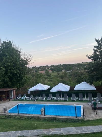 The image depicts an outdoor swimming pool area at dusk in General Kiselovo, Bulgaria. The pool is rectangular with bright blue water and is surrounded by a black metal fence. A lawn with green grass extends in front of the pool, separated by a stone pathway. Behind the pool, there are several lounge chairs and white umbrellas, some displaying Coca-Cola logos and one with the word "БАНКЯ" (Bankya, a Bulgarian town). To the left, a small brick structure with a bar and stools is visible. In the background, a hillside village with scattered houses and lush green trees is nestled under a pale pink and blue sky, with faint contrails from an airplane visible. A person in a green t-shirt and black shorts is walking away from the camera near the edge of the pool.