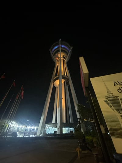 The image captures a night scene in Alor Setar, Malaysia, featuring the Alor Setar Tower. The tower, a prominent landmark, is illuminated with blue lights at its apex, creating a striking contrast against the dark sky. Several slender white beams radiate outwards from the upper section of the tower, supporting its distinctive saucer-shaped top.

In the foreground and extending towards the bottom of the frame, a series of poles with small red flags are visible. These flags appear to be part of a procession or celebration. In the upper portion of the image, a large sign is partially visible, oriented sideways. Text on the sign includes "Abdy" and "Dj." The sign seems to be advertising or providing information related to the tower or an event.

The overall atmosphere is nocturnal, with artificial lighting playing a key role in highlighting the architectural features of the tower and creating a festive or celebratory mood suggested by the flags. The presence of trees and other background structures indicates an urban setting. The image is shot from a low angle, emphasizing the height and grandeur of the Alor Setar Tower.