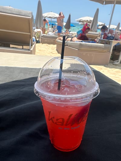 A close-up shot of a red beverage in a clear plastic cup with a dome lid and black straw, resting on a black surface. The cup has a logo that reads "Kalo" and "on the beach" in faint lettering. In the background, a busy beach scene unfolds. Several beach chairs are visible, some occupied by people. Umbrellas provide shade, and the sandy beach meets the turquoise sea under a clear blue sky. People in swimwear are scattered across the beach and in the shallow water. The overall atmosphere suggests a sunny day at a resort in Ayia Napa, Cyprus, known for its vibrant beach culture.