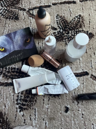 A flat lay of assorted cosmetic products is arranged on a textured, beige and brown rug. The items include a light-colored foundation bottle with a black cap and graphic design, a small glass bottle with a white dropper lid, a rose gold colored lipstick or balm, a light brown makeup sponge, two white squeeze tubes, a white stick deodorant, and a small tube of ointment. One of the squeeze tubes has the brand name "Eno" printed on it, and the other has text in Russian. The white stick deodorant is labeled "Perfect Balance 9:1 Matcha Airy Fit Sun Stick." The background rug has a pattern of dark brown and black abstract shapes against a lighter beige. The overall impression is a collection of beauty products laid out casually. No people are visible in the image, and no specific location in Ташлак, Kyrgyzstan, can be determined from the scene.