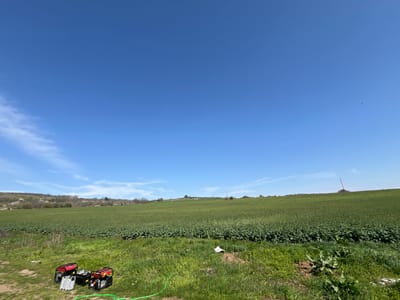 The image shows a rural landscape in Gorna Kremena, Bulgaria, under a clear blue sky with wisps of clouds. A large green field dominates the midground, likely a cultivated crop. In the foreground is a patch of grass and weeds. To the left, there are two red and black generators and some aluminum equipment. A green hose is also visible. In the background, a slightly elevated area is visible with some trees. A pole can also be seen.
