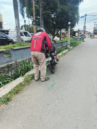 A person in a red and black "PegiLagi" jacket ties green string to the back of a parked motorcycle. The license plate reads "5799 FX." The motorcycle sits on the side of a road next to a curb and a ditch. The person wears tan pants, sandals, and a helmet. Trees, utility poles, and buildings are visible in the background.