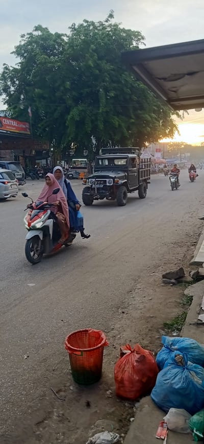 This media file depicts a street scene in Gampongteungoh, Indonesia, during what appears to be sunset or early evening. The sky is hazy with the sun low on the horizon, casting a warm glow. The overall atmosphere suggests a bustling, everyday environment.
The primary subjects are people on motorcycles. In the foreground, two individuals are riding a scooter. The passenger, wearing a pink hijab, is looking towards the camera. The driver, wearing a blue hijab and a blue top, is holding a blue plastic bag. Further down the road, other motorcyclists are visible, with one person in the distance wearing a red and white outfit and another in a light-colored hijab.
A significant object in the scene is a vintage black Jeep-style vehicle with a cargo bed, its tires large and rugged. It is positioned on the dirt road, with a person driving. In the background, there are various buildings and structures, including one with a sign that reads "CENTRAL coffee" in red lettering, suggesting a commercial area.
The road is unpaved and dusty, indicative of rural or semi-urban Indonesian settings. On the side of the road, there are several trash receptacles and bags of refuse. An orange plastic basket is filled with what appears to be green waste, and several large blue plastic bags and one red plastic bag filled with trash are also present.
The time of day is clearly indicated by the warm, diffused sunlight and the lengthening shadows. The weather appears to be clear but with some atmospheric haze. The location context of Gampongteungoh, Indonesia, is provided. The scene captures a moment of daily life, with people commuting and the mundane reality of waste management alongside commercial activity. There are no overtly strong emotions conveyed, but rather a sense of ordinary activity.