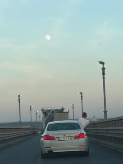 The image depicts a white BMW wagon on a bridge, likely a highway, with a person leaning over the trunk of the car. The person is wearing a light-colored shirt. The car has a license plate that reads "SV 55 WWM". In the background, there are streetlights lining the bridge. The sky is light blue with some wispy clouds, and the moon is visible as a bright disc. The scene appears to be set during the daytime, possibly late afternoon or early evening, given the soft light. The overall impression is that of a roadside stop or a pause on a journey, with the bridge and the distant landscape suggesting an open-road setting. The specific location is identified as Giurgiu, Romania.