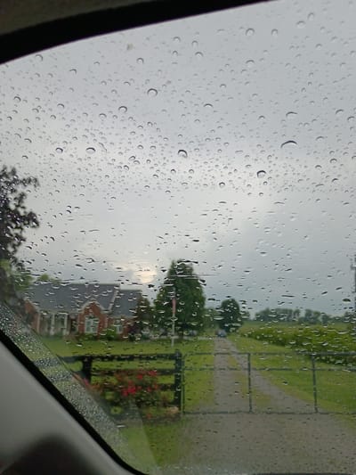 The image is a photograph taken from inside a vehicle, looking out through a rain-streaked windshield at a rural or suburban landscape. The foreground shows the edge of the vehicle's interior. The middle ground features a house with red brick and white trim, a black wooden fence, and a patch of red flowers. A gravel driveway leads away from the house towards a gate and a wider road in the distance. The background consists of green fields, trees, and an overcast sky. The scene is set in Bagdad, United States.