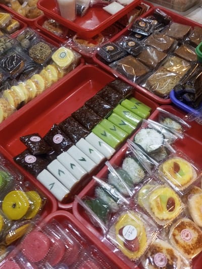 This image displays a high-angle view of a meticulously arranged assortment of individual snack portions, likely for sale, located in Lê Thanh Nghị Ward, Vietnam. The scene is dominated by multiple red plastic trays, each containing various types of sweets, cakes, and pastries, all individually packaged in clear plastic wrap, bags, or small transparent containers.
Prominent items include:
* Rows of dark brown, glossy, rectangular blocks, likely a type of sticky rice cake (kueh or dodol), individually wrapped in clear film.
* Adjacent to these are rows of light green rectangular blocks, also individually wrapped, and white rectangular blocks featuring a green 'V' or leaf design.
* Numerous small, round, yellowish tarts or pies, some topped with fruit or jam, are presented in clear plastic cups with paper liners. Several of these tarts display small, circular, brand-specific stickers.
* Slices of yellowish rolled cake, often resembling a Swiss roll, are individually wrapped in clear plastic.
* Small, round, white items, possibly rice cakes or glutinous rice balls, some coated in shredded coconut, are contained within clear plastic bags.
* Various square or rectangular plain cakes and loaves, appearing dense and brownish, are also visible in clear plastic packaging.
* Towards the upper left, smaller pastries and buns are displayed. In the upper center, small cups or pouches contain light pink and white liquids, also individually sealed.
* In the lower right, multiple small, round, browned items resembling mini pancakes or fritters are present, some marked with pink circular stickers.
No individuals or active interactions are visible. The items are presented in an organized manner consistent with a retail food display or market stall.