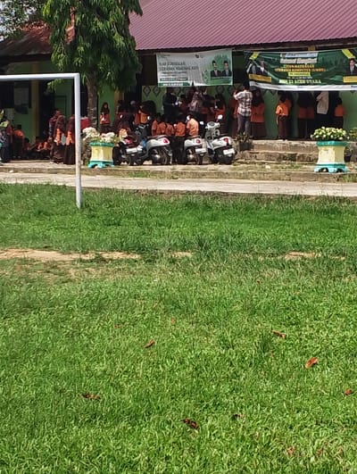 The image depicts a scene outside a building, likely a school, in Pantonrayeuk Satu, Indonesia. Several people, including children in school uniforms (orange shirts and brown skirts/pants) and adults, are gathered on steps and in front of the building. Some are standing, while others are sitting on the ground.
Two motorbikes are parked on the steps, one white and one dark colored. A large tree with green foliage is visible to the left of the building. Banners with text and images of men are displayed on the building's facade. A white pole, part of a structure, is prominent on the left side of the image.
In the foreground, a well-maintained grassy area with some fallen leaves is present. The overall impression is of an outdoor event or gathering at an educational institution. The building appears to have a red-tiled roof.