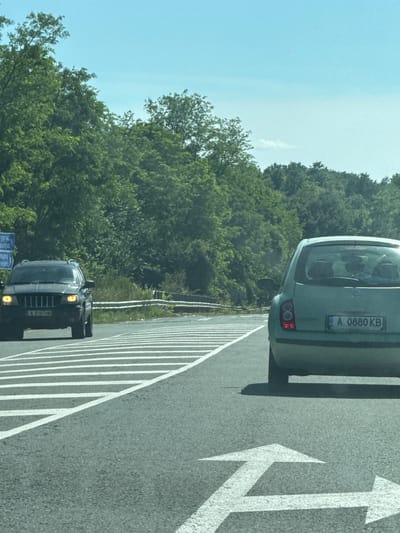 The image shows a two-lane highway with dense green trees lining the sides. Two cars are visible on the road. In the left lane, a dark colored Jeep Grand Cherokee is approaching the camera. Its headlights are on, and the license plate is partially visible. In the right lane, a light green hatchback, possibly a Nissan Micra, is moving away from the camera. Its license plate reads "A 0880 KB". Road markings, including directional arrows and lane dividers, are clearly visible on the asphalt. A blue road sign with Cyrillic text is present on the left side of the road, indicating "Burgas". The sky is clear and blue with some faint clouds. The scene appears to be a sunny day on a highway in or near Drachevo, Bulgaria, given the signage.