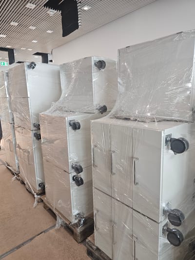 The image shows a row of large, white cabinets, each covered in clear plastic wrap. The cabinets appear to be modular and have silver handles and black casters. They are placed on wooden pallets on a concrete floor. In the background, a ceiling with a slatted design and lights is visible, along with a green exit sign. The scene suggests that these cabinets are either being stored, moved, or prepared for installation. The setting appears to be an indoor space, possibly a warehouse, storage facility, or a room under construction or renovation within the city of Darmstadt, Germany. There are no people visible in the image.