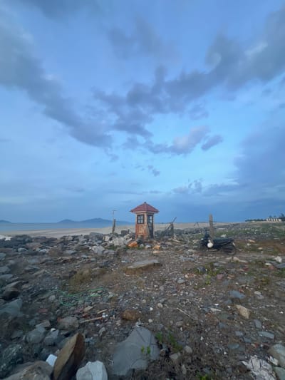 The image depicts a desolate beach scene, likely near Hoi An, Vietnam, characterized by a rocky shoreline and a cloudy sky. A small, weathered wooden structure with a red-tiled roof and a single window stands prominently in the foreground, resembling a guard post or a small shelter. In the middle ground, a black scooter with a white helmet is parked, and a wooden post with some equipment is visible. The beach itself is strewn with rocks of various sizes, some debris, and sparse vegetation. The sea stretches out to the horizon, with distant mountains visible under a heavily clouded sky, suggesting either early morning or late evening light. The overall impression is one of a remote and somewhat neglected coastal area.
