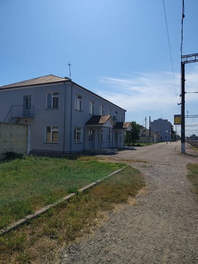 The image depicts a modest, two-story, light blue building situated next to a railway track. The building appears to be a station or administrative facility, featuring several windows and a small covered entrance area. To the left of the building, there is a low concrete wall and a patch of green grass. A gravel path leads away from the building towards the railway line, which is visible in the background along with overhead electrical infrastructure typical of a train station. The sky is clear and blue with a few wispy clouds. A street light and a yellow sign with text, likely informational, are also visible near the railway platform. The scene suggests a sunny day in a semi-urban or rural setting. The location is identified as Bakhmach, Ukraine. There are no people visible in the image.
