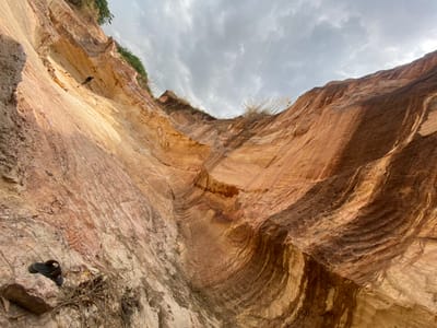 This image, captured in Amangwu, Nigeria, presents an upward perspective of a deep, excavated or naturally eroded geological formation. The scene is dominated by towering, stratified earthen walls composed of reddish-brown, orange, and light beige sediments. Prominent vertical striations and undulating horizontal patterns are visible across the wall surfaces, indicative of erosion or excavation. An overcast, cloudy sky is visible at the top. At the lower-left, a single dark-colored sandal rests on a rock. High on the left wall, a small, dark object resembling a bird is clinging to the surface. Sparse dry vegetation is present along the upper ledges and scattered near the base of the walls. No human figures are present.