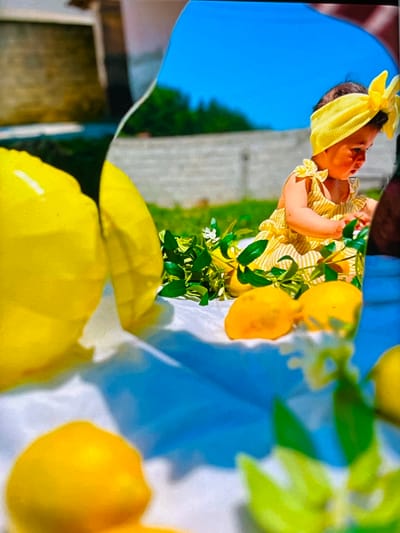 This media file shows a close-up, rotated view of a baby wearing a yellow headband and a yellow striped outfit. The baby is looking upwards and to the right. In the background, there are several lemons on a branch with green leaves and white flowers. The setting appears to be outdoors, with a bright blue sky visible on the left side of the image, and greenery suggesting trees or bushes. The lighting suggests it is daytime, possibly sunny, given the vibrant colors. The overall mood is cheerful and summery, evoked by the bright yellow theme and the baby's presence. The image is somewhat abstract due to the close-up and rotated perspective, focusing on textures and colors rather than a clear, wide view of the scene. There is no visible text in the media file.