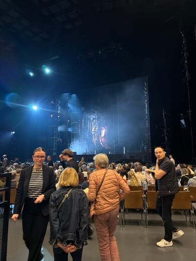 The image depicts an indoor performance venue in Boulogne-Billancourt, France. The scene captures audience members within a darkened hall, orienting towards a brightly lit stage in the background. The stage features a complex set design with large dark panels, scaffolding-like structures, and a central element displaying an abstract or structural motif. Multiple bright blue stage lights illuminate the stage and atmospheric haze. In the immediate foreground, four individuals are prominent: A woman with dark hair and glasses, wearing a black blazer over a black and white striped shirt and black pants, walks left. Adjacent to her, a blonde-haired woman, viewed from behind, stands wearing a dark blue denim jacket. To her right, an older woman with short grey hair, also seen from behind, walks right in a light peach-colored puffer jacket and matching pants. On the far right, a man with dark hair, attired in a black short-sleeved shirt, black vest, black pants, and white sneakers, stands facing right with his left hand near his ear. Multiple rows of brown upholstered seats, occupied by numerous additional audience members, extend into the mid-ground and background, all facing the stage. The ceiling is dark, revealing overhead lighting rigs.