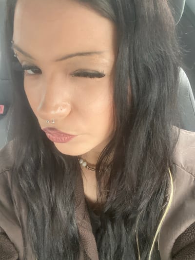 A female individual is depicted in a close-up portrait, winking with her right eye while her left eye remains open. She has long, dark hair, with a single light-colored highlight visible on the right side of the frame. Facial piercings include a silver barbell in her left eyebrow and a silver horseshoe ring in her septum. Her lips are pursed and covered with dark red lipstick. She wears a dark brown zippered jacket, consistent with fleece material, and a beaded necklace featuring alternating light and dark beads around her neck. The background is out of focus, revealing the dark upholstery of a vehicle seat. This scene is located in Taverny, France.