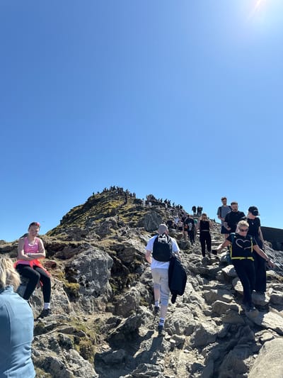 This image depicts a crowded mountainside with numerous people climbing and resting. The location is in Beddgelert, United Kingdom, evidenced by the rocky terrain and the presence of hikers in typical outdoor attire. The sky is clear and bright blue. People of varying ages are seen scattered across the rocky slope, some moving upwards and others pausing. The landscape is characterized by large, grey rocks and sparse vegetation. Some individuals are carrying backpacks.