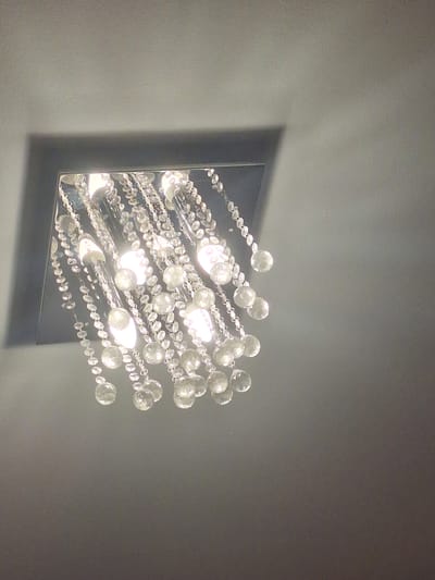 The image shows a close-up of a modern chandelier hanging from a ceiling. The chandelier features a square, reflective base and is adorned with numerous strands of faceted crystal beads and small, bright light bulbs. The crystals catch and reflect the light, creating a sparkling effect. The chandelier is positioned against a plain, textured gray ceiling, and a subtle shadow cast by the chandelier's base is visible on the ceiling to the left. The lighting in the image is somewhat diffused, suggesting it might be daytime, or the room's ambient lighting is dim, allowing the chandelier to be the primary light source. The overall mood is elegant and contemporary. There is no visible text in the image. The context provided indicates the location is Mińsk Mazowiecki, Poland.