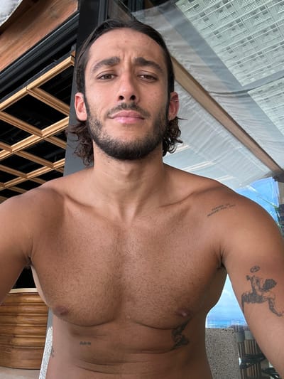 A shirtless man with curly brown hair and a beard faces the camera. He has multiple tattoos on his arms and chest. The background includes a window with a textured design and what appears to be a tiled surface. The photograph has been taken at a tilted angle.