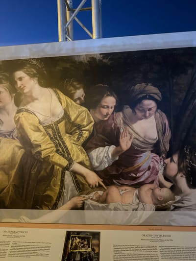 The image displays a section of a large painting, framed by a metal structure and a dark blue sky, suggesting it is outdoors. The painting depicts a biblical scene with several women gathered around a baby. The central focus is a baby lying on its back, wrapped in white cloth. To the left, a woman in a shimmering gold dress, adorned with dark, intricate trim and pearls, gazes down at the baby, with her left hand pointing towards it. Behind her, another woman in a mauve dress with a draped neckline also looks at the baby. To the right, two other women are partially visible. One in a dark head covering leans in close, her face obscured, while another woman with dark hair and a bejeweled headdress is seen in profile. The background of the painting is dark and appears to be a landscape with foliage. Below the painting is a display board with text, including titles like "PINTURA ITALIANA" and "ORAZIO GENTILESCHI," and descriptions of the artwork. The text mentions "Mazarrón, Spain," suggesting the painting is displayed or exhibited in this city.