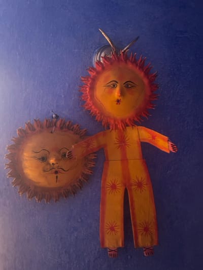 Two sun-themed ceiling decorations adorn a blue surface. The upper one has a painted face and a body-shaped appendage extending outward, resembling a person in a suit, decorated with orange stars. The lower sun-like figure also has a painted face but without body parts. A lighting fixture is positioned beside the first sun. The decorations appear handmade.