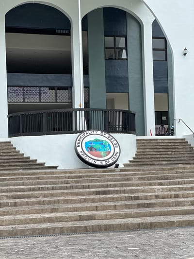 The image shows the exterior of a municipal building in Oslob, Cebu, Philippines. In the foreground, a wide, stone staircase leads up to the building's entrance. At the base of the stairs, a large circular seal is prominently displayed, reading "Municipality of Oslob, Province of Cebu." The seal features a landscape with a sailboat on water, trees, and mountains under a sky with stars and a sun. The building itself is modern with large arched entrances and teal and white painted sections. A black metal railing curves around a balcony area on the second level. A portion of a sign with text, including "Rights First Legislation Forward Strategic Action for Equal Participation and Inclusive Development," is visible through a window on the second floor. A motorcycle is parked on the right side of the building. The sky appears overcast, and there are visible raindrops, suggesting it may be raining or has recently rained.