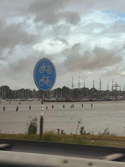 The image shows a blue circular sign with two white bicycle symbols, indicating a bicycle path, mounted on a pole. The sign is in the foreground, slightly out of focus. In the background, there is a body of water, likely a harbor, with numerous sailboats docked. Trees line the far shore, and the sky is overcast with dramatic, cloudy formations. The scene appears to be from a coastal area, possibly near a marina in Enkhuizen, Netherlands, given the prevalence of sailboats and the style of the bicycle sign. The water reflects the cloudy sky, creating a muted, grey-blue hue. There are several dark posts or buoys visible in the water, marking the edge of a channel or mooring area. A portion of a road or pathway is visible in the bottom of the frame, suggesting the perspective is from a moving vehicle or a roadside vantage point. The overall atmosphere is calm and somewhat subdued due to the cloudy weather.