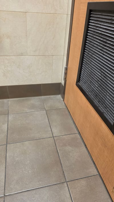 The image shows a close-up view of the corner of a room with tiled walls and floor, and a wooden door with a vent. The wall tiles are beige and rectangular, laid in a horizontal pattern. The floor tiles are square and a light brown or taupe color, with dark grout lines. A dark brown baseboard runs along the bottom of the wall. To the right of the wall is a wooden door, with a darker brown frame around a rectangular vent that has horizontal slats. The door appears to be hinged on the left side from the perspective of the image. There are no people or animals visible in the image. The scene is indoors, and the lighting suggests it is a well-lit interior space. The location of Haughton, United States of America is not visually discernible from the image itself; this information is provided externally.