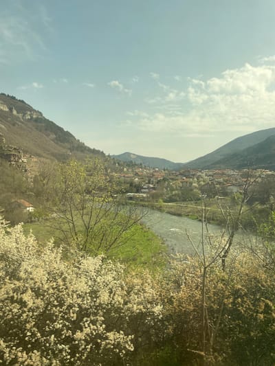 The image shows a landscape in Tserovo, Bulgaria, featuring a river flowing through a valley. On the left side of the river, there is a hillside with a mountain peak towering above. A small town is nestled between the hillside and the river. The right side of the riverbank is covered in lush greenery and trees with white blossoms. The sky is partially cloudy with a light blue hue. There are no people visible in the image.