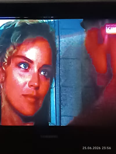 This is a close-up, pixelated image of a woman's face, seemingly captured from a screen, possibly a television or computer monitor, given the visible date and time stamp in the bottom right corner. The woman's face is heavily saturated with red, giving her skin a somewhat unnatural glow. Her eyes are blue and appear to be looking slightly off-camera. Her hair is curly and styled back from her face.

To her right, a large, abstract shape in a bright red hue dominates the right side of the frame. Partially visible within this red shape, and also in the top right corner of the image, is a bright white and pink logo or text that reads "KIN..." suggesting a movie or entertainment context. The background to the left of the woman's face is a blurry, textured blue, possibly representing a wall or a scene element. The overall impression is that of a paused or distorted video frame.

The date and time stamp at the bottom reads "25.06.2026 23:56". The location context is given as Bolhrad, Ukraine, but there are no specific visual cues in the image to confirm this location. The overall mood is difficult to ascertain due to the image distortion and heavy color filtering, but the woman's expression is neutral.