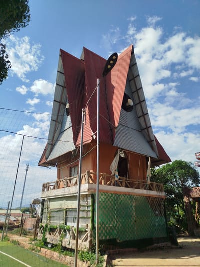 A multi-story building with a distinctive roof stands in An Phu Ward, Vietnam, under a partly cloudy sky. The building features a layered roof design with red and gray corrugated metal. The building is orange and includes a balcony with faux-stone railings. The base of the building has green, diamond-patterned panels. A tall metal light pole stands in front of the building. To the left is a green field enclosed by a net fence. Other buildings and trees are visible in the background.