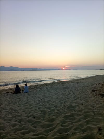 The image captures a serene sunset scene on a sandy beach in Naxos, Greece. Two individuals, one in a dark shirt and the other in a light-colored shirt, are seated on the sand, facing the calm sea. The sun is partially visible on the horizon, casting a warm, orange glow across the water and the sky, which transitions from a pale blue at the top to a soft yellow and pink near the horizon. Distant mountains form a silhouette against the vibrant sky. The beach sand is textured with ripples and a few scattered pebbles. The gentle waves of the Aegean Sea lap at the shore. The overall atmosphere is peaceful and contemplative.