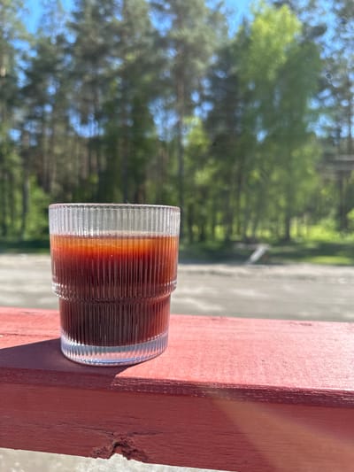 The image shows a glass of dark liquid on a red wooden surface, possibly a ledge or windowsill. The glass has a clear, ribbed design. The liquid appears to be layered with varying shades of brown. In the background, blurred trees and sky are visible, suggesting an outdoor setting. The presence of trees indicates a natural environment or a park-like area. There is no explicit information in the image indicating the location is Askat, Russia, or about any people, actions, or interactions.