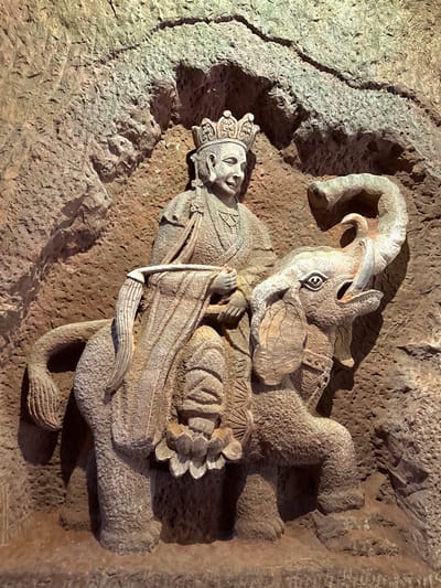 This is a detailed stone relief carving embedded into a rough, textured rock wall, likely within a grotto or cave setting in Shizhong, China. The carving features two main subjects: a crowned human figure and a mythical beast.

The human figure, possibly a deity or royalty, is depicted in a reclining or lying posture. They wear an elaborate crown with intricate patterns and appear to be dressed in layered robes, intricately carved with folds and details. The figure holds a long, slender object resembling a staff or sword diagonally across their body. Their facial expression is serene and calm.

Positioned above and partially behind the human figure is a large, mythical creature. This beast has prominent horns curling backward, large, stylized eyes with a distinct pupil design, and a broad snout. Its body is covered in textured scales, and a tortoise-like shell is visible on its back. The creature's body curves and wraps around the human figure, creating a dynamic composition.

The entire carving is rendered in a monochromatic stone, with variations in shade and highlight brought out by strong, directional lighting that casts deep shadows, enhancing the three-dimensional quality of the relief. The surrounding rock wall is rugged and natural, suggesting the carving is an integral part of the geological formation. There is no visible text in the image.
