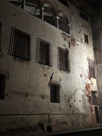 The image shows the exterior of a dilapidated old building, possibly a historical site, at night. The facade is made of weathered plaster over brick, with significant areas of peeling paint and exposed brickwork, indicating disrepair. Several windows are visible, many covered with heavy, dark metal bars, suggesting a historical or security purpose. Some windows are dark, while others show faint light or reflections from within.
The architecture includes arched openings and columns on an upper level, creating a cloistered or loggia-like effect. The lower part of the building features more visible brickwork and what appear to be doorways or darkened openings. The lighting is dim and atmospheric, with shadows cast by the architectural features and the night sky. The overall impression is one of age, decay, and historical significance.
This building is located in Lagnasco, Italy, and is identified as the "Torre Tonda" or "Rotonda", a historical tower that is part of the Lagnasco Castle complex. The Torre Tonda is notable for its unique circular shape and its history as a defensive structure and later as a prison. The barred windows are consistent with its former use as a jail.