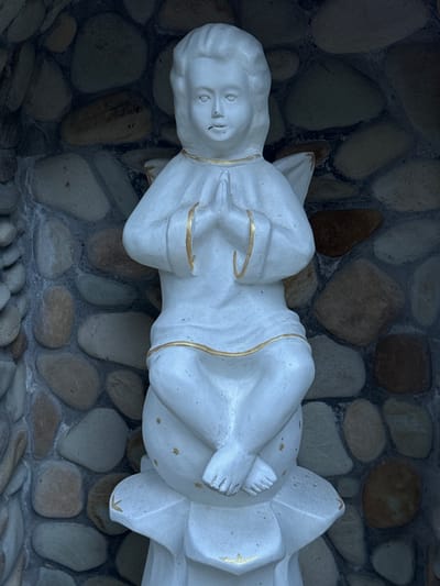 A white statue of a cherub with gold trim is shown from the chest up, sitting cross-legged on a lotus-shaped base. The statue is positioned in an alcove with a stone wall behind it. The stone wall is made of various-sized, irregularly shaped river rocks in shades of grey, brown, and tan, some of which are smooth and rounded, while others are more angular. The statue's hands are clasped in front of its chest, and it has feathered wings extending from its back. The cherub has a youthful face with closed eyes, a serene expression, and curly hair. The overall lighting suggests it may be daytime, but the alcove is somewhat shaded. The image is cropped to focus on the statue and its immediate surroundings, with no other people or objects visible. The location is identified as Lisnyky, Ukraine.