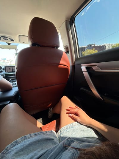 The image depicts the interior of a modern passenger vehicle, viewed from a low angle in the rear seat, looking towards the front and slightly right. In the foreground, the bare legs and lower torso of a person are visible, wearing light-colored denim shorts. Their right hand, adorned with a ring on the ring finger and manicured nails, rests on their right thigh. The back of the front passenger seat, upholstered in reddish-brown leather with visible stitching, occupies the central portion of the frame. The headrest of this seat is also reddish-brown. To the left, the lower leg and foot, wearing light-colored sneakers, of a second individual are visible in the front passenger footwell. The dashboard and a digital infotainment screen are partially visible through the gap between the front seats. The right rear door panel, dark-colored with a distinctive metallic silver, angled door handle and horizontal trim, is prominent on the right. Through the car windows, a clear blue sky is visible above a distant urban or suburban landscape. Buildings with varying architectural styles, including one with a tiered roof structure, and green and yellowish tree foliage are present. Thin overhead power lines intersect the sky. The car's headliner is light-colored, and an overhead console with buttons, a rearview mirror, and a small hanging accessory are visible near the windshield. Bright direct sunlight illuminates the interior, casting strong shadows. The scene is consistent with an urban environment, such as the city of Novoivanovskoe, Russia.