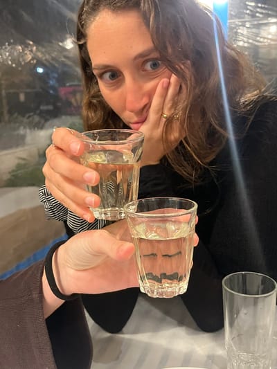 This image captures two individuals at a table within an establishment in Megalochori, Greece. A woman with wavy brown hair, wearing a black long-sleeved top, occupies the upper portion of the frame. Her left hand extends forward, holding a small, clear, faceted glass containing a translucent liquid. Her right hand rests against her face, and she looks directly at the camera with wide eyes and slightly pursed lips. A gold ring is visible on her right ring finger, and a black and white striped scrunchie is on her left wrist. The left hand and forearm of a second individual, clad in a dark brown long-sleeved garment and a black wristband, are visible in the lower left, holding an identical clear glass with translucent liquid. An empty matching glass rests on the white tabletop to the lower right. The background features blurred lights observed through a transparent enclosure, indicating an indoor or enclosed outdoor setting, likely during nighttime.