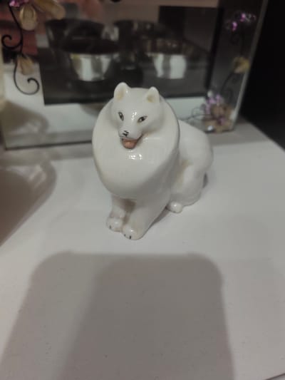 A white ceramic figurine of a fluffy dog sits on a light-colored surface. The dog is positioned in the center of the frame, facing slightly to the left. It has large, expressive eyes, a small black nose, and an open mouth revealing a pink interior. Its body is rounded, and its paws are visible at the front. The background is blurred, showing a reflective surface with some decorative elements, suggesting it might be on a shelf or table. The lighting casts a shadow of the figurine onto the surface in front of it. The image does not provide any information or visual cues related to Ostafievo, Russia.