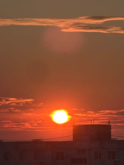 The image captures a sunset scene over a city, presumably Bakhchysarai, Russia. The sky is a vibrant gradient of orange and red, with wispy clouds tinged with the same warm hues. The sun, a bright, glowing orb, is prominently positioned in the lower-middle section of the frame, casting its light across the sky. In the foreground, the silhouette of buildings with flat roofs and visible window frames is discernible. Some electrical wires crisscross the scene. The overall impression is one of a tranquil evening, with the sun setting behind the urban landscape.