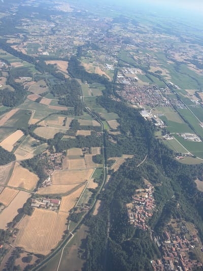 This is an aerial photograph taken from a high altitude, likely from an airplane. The image shows a vast agricultural landscape in Pombia, Italy, with a mix of cultivated fields, forests, and scattered settlements. The sun appears to be high in the sky, suggesting it is midday. The weather seems clear, with good visibility.

The dominant subjects are the agricultural fields, which are varied in color and texture, indicating different crops and stages of cultivation. Some fields show straight rows of crops, while others are covered in what appears to be harvested hay, bundled into small, round bales. Patches of dense green forest are interspersed throughout the landscape, creating a striking contrast with the lighter tones of the fields.

Several small towns and hamlets are visible, characterized by clusters of red-tiled roofs. These settlements are nestled within the agricultural land and surrounded by trees. One notable settlement is situated on a slightly elevated area, surrounded by greenery. A man-made waterway, likely a canal, runs through the scene, bordered by trees. Roads and smaller paths crisscross the landscape, connecting the settlements and fields.

There are no visible people or specific objects that stand out in terms of human activity, other than the general signs of agricultural production and the presence of human settlements. The overall scene depicts a rural, European countryside bathed in bright sunlight. There is no visible text in the image.
