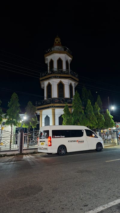 The image depicts a night scene in front of a mosque minaret in Lhokdagang, Indonesia. A white passenger van with the markings "PT. MUTIARA EXPRESS" and "Kembar Group" is parked on the right side of the street, facing left. The van has its brake lights illuminated. Several evergreen trees are planted between the van and the mosque. The mosque minaret is octagonal and illuminated, featuring gold and white decorative elements. Power lines are visible against the dark night sky. The street in the foreground is asphalt with white lane markings. No people are visible in the image.